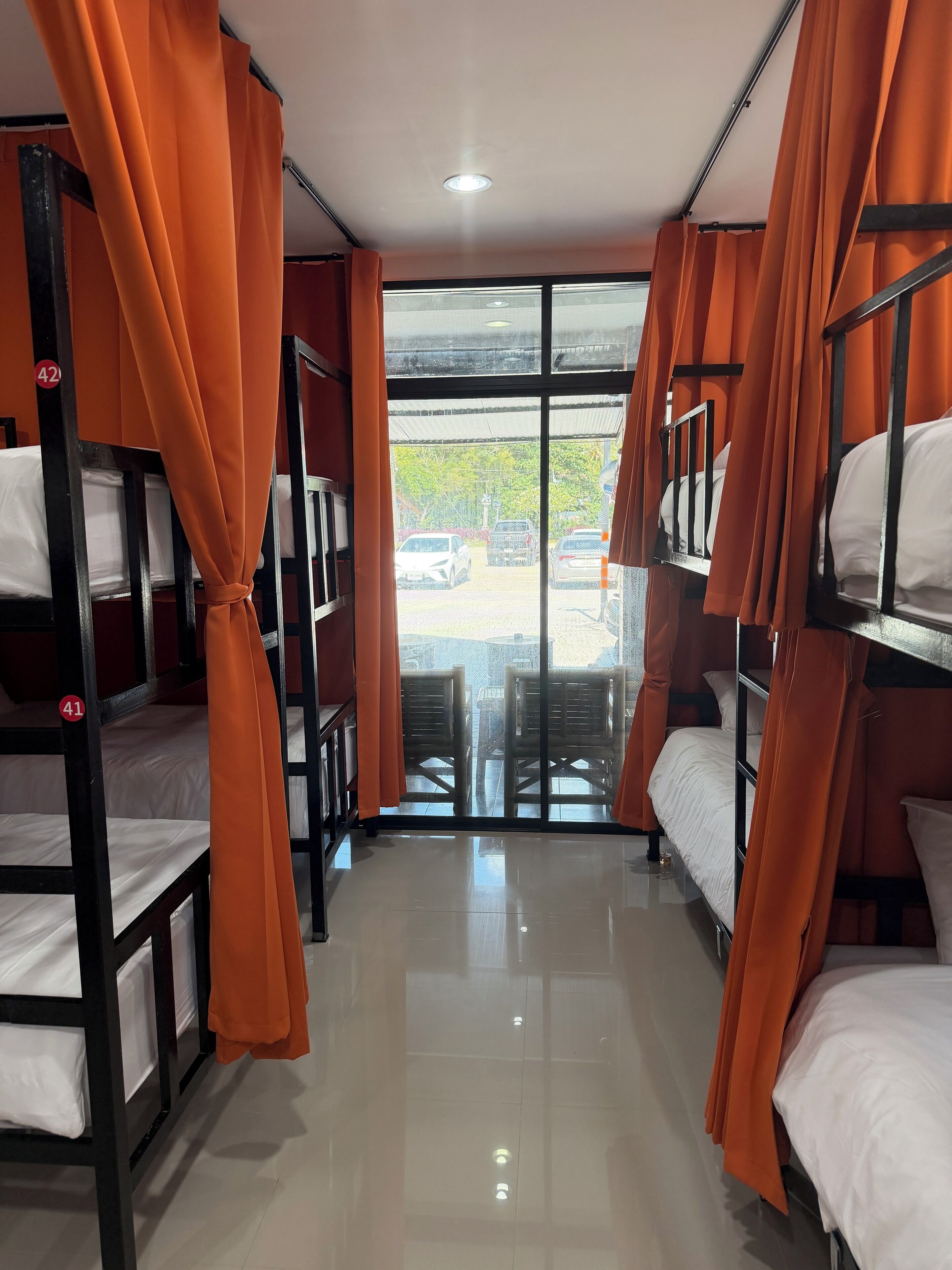 A room with bunk beds, orange curtains, and a view of parked cars outside.