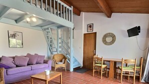 Un soggiorno con un divano viola, un tavolino in legno, una poltrona in vimini, un tavolo da pranzo con sedie, una scala e una porta.