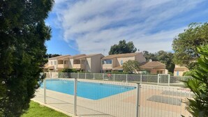 A swimming pool with a metal fence, surrounded by greenery and residential buildings in the background. - Joli Mazet T3 avec piscine (La Londe-les-Maures)