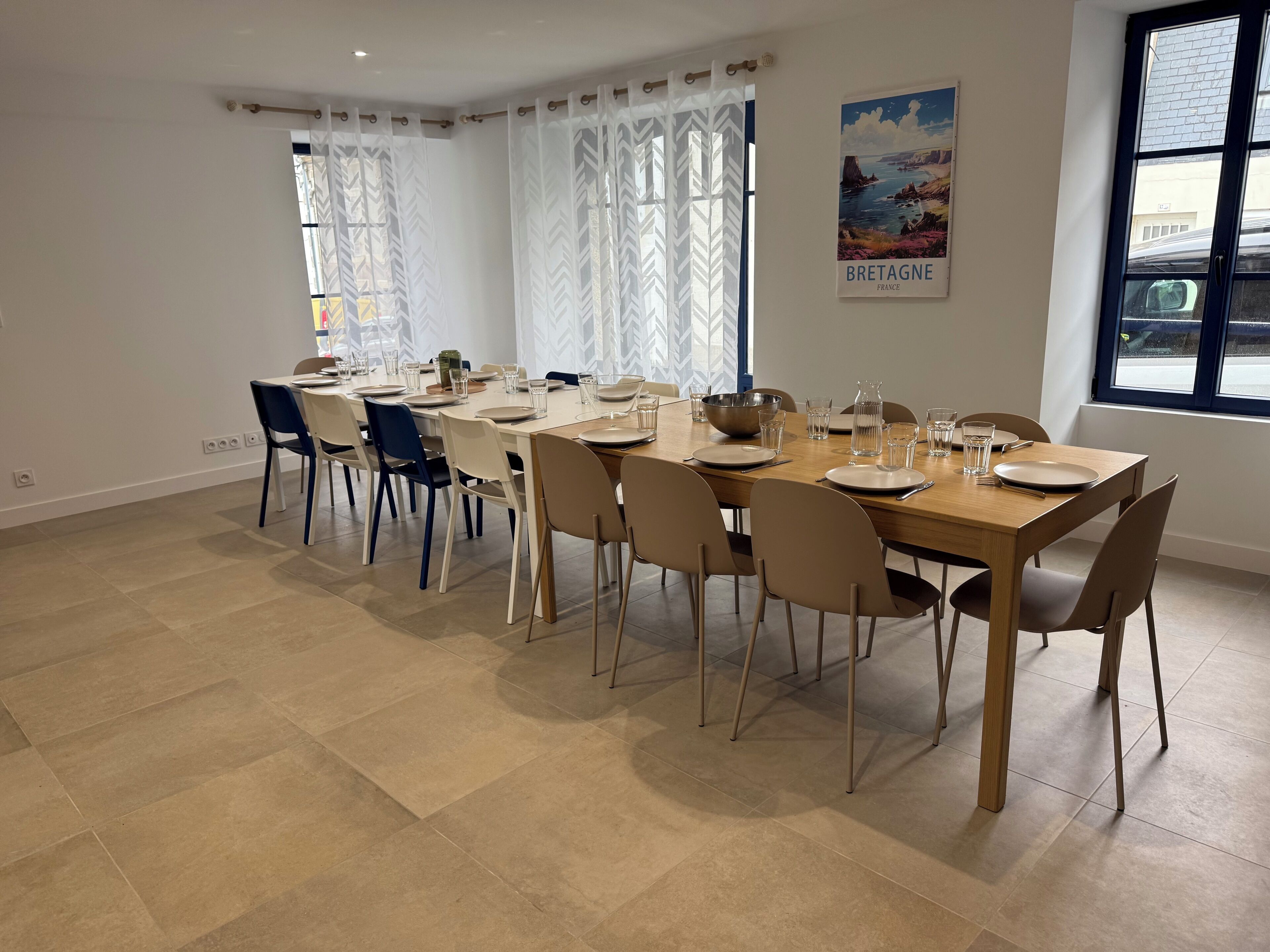 A dining area with a long table set for six, surrounded by chairs, in a room with a large window and a poster of Bretagne, France.