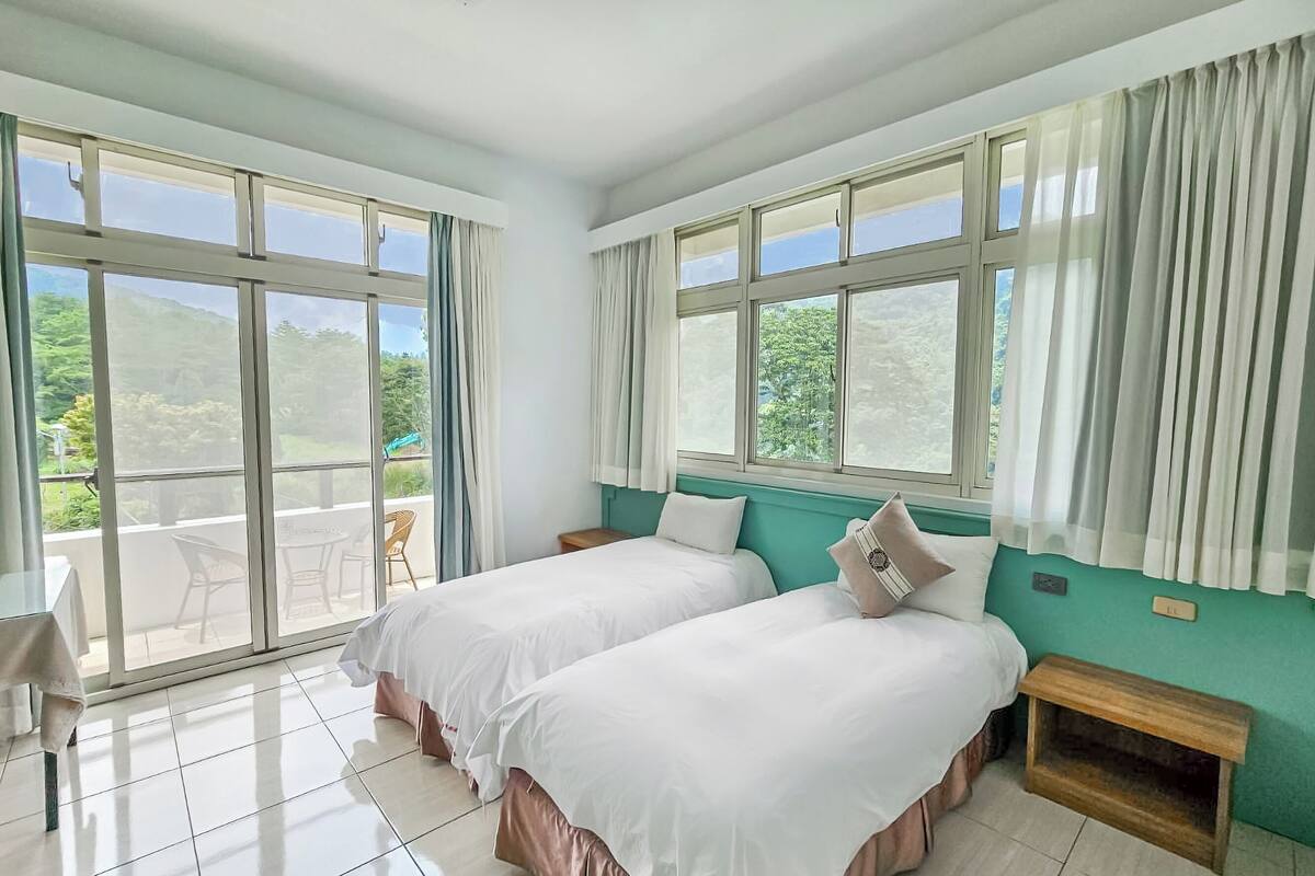 Une chambre d’hôtel avec deux lits, un balcon avec des chaises et de grandes fenêtres avec des rideaux.