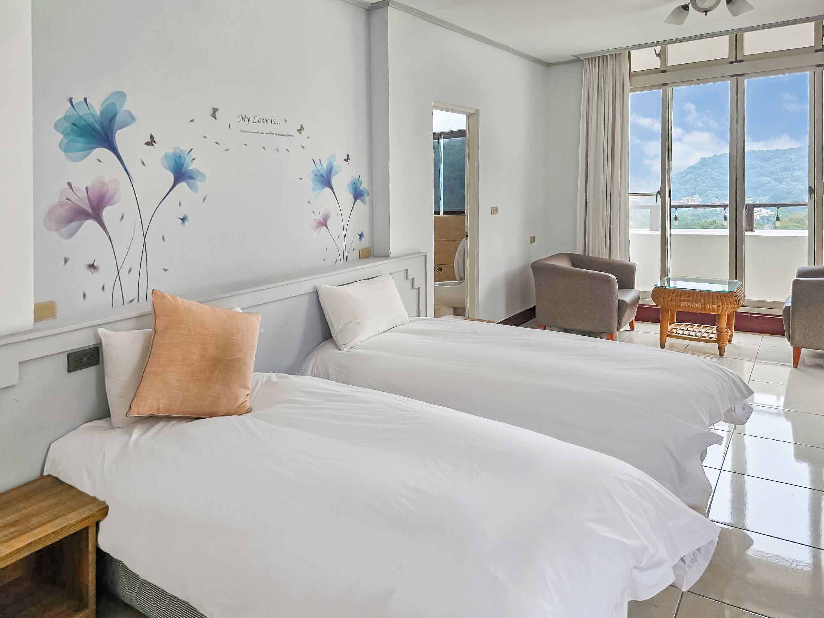 Une chambre d’hôtel avec deux lits, une petite table, une chaise et une grande fenêtre donnant sur l’extérieur.