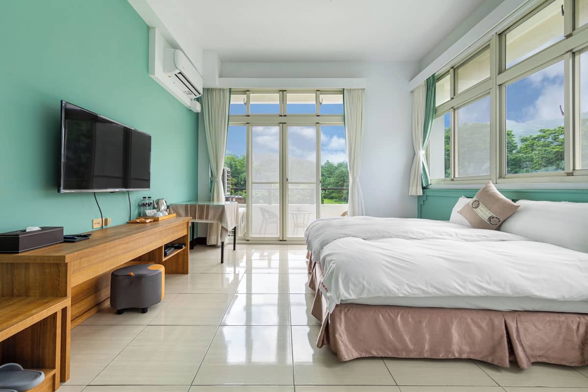 Une chambre d’hôtel moderne dotée d’un grand lit, d’un bureau en bois, d’une télévision à écran plat et d’un balcon offrant une vue sur la verdure.