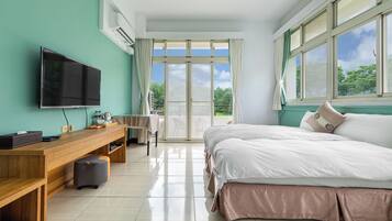 A modern hotel room with a large bed, a wooden desk, a flat-screen TV, and a balcony with a view of greenery.