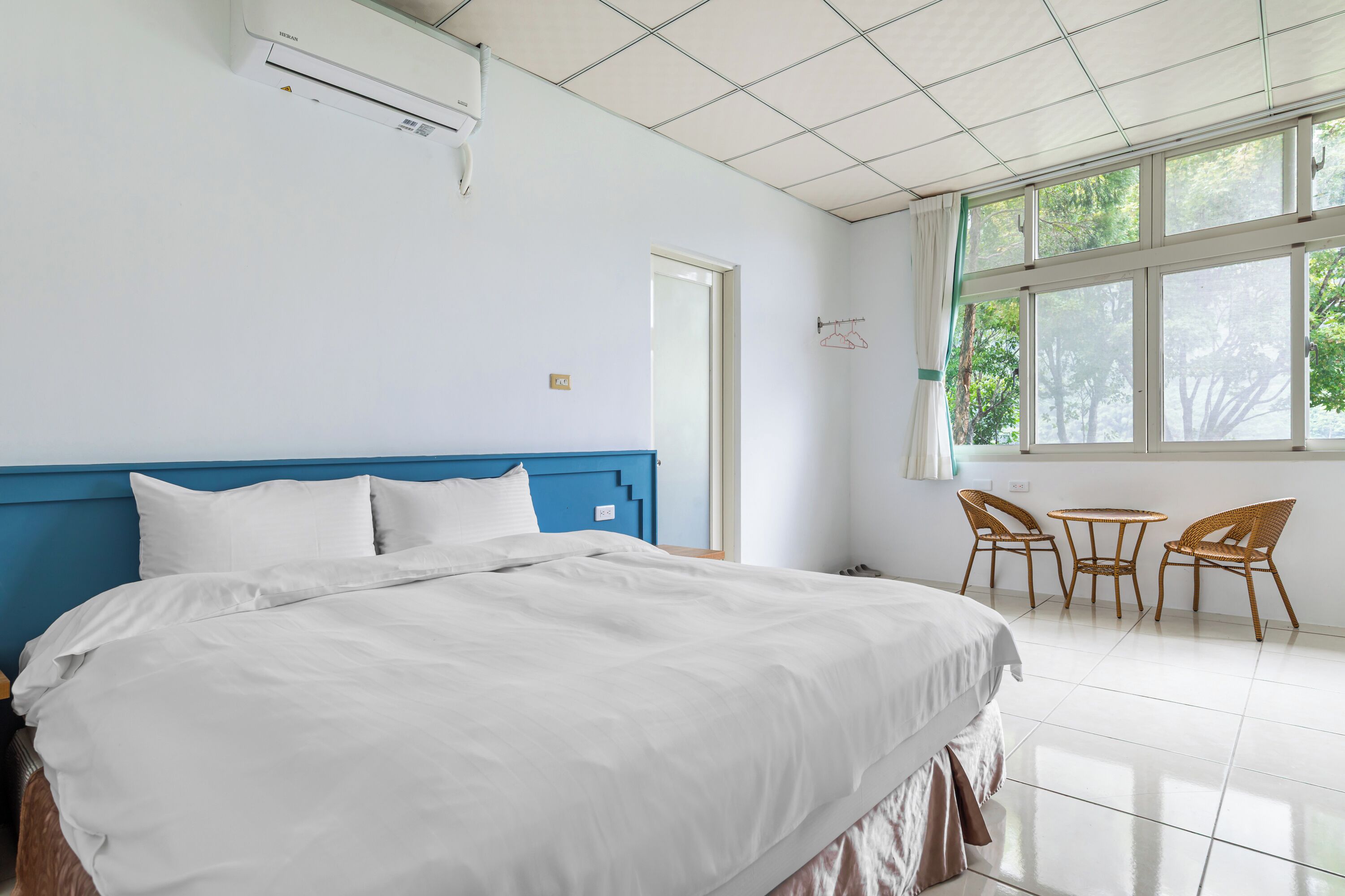 Habitación de hotel con una cama grande, cabecera azul, una pequeña mesa con sillas y ventana con vistas a árboles.