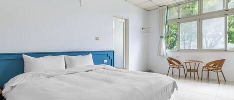 Habitación de hotel con una cama grande, cabecera azul, una pequeña mesa con sillas y ventana con vistas a árboles.