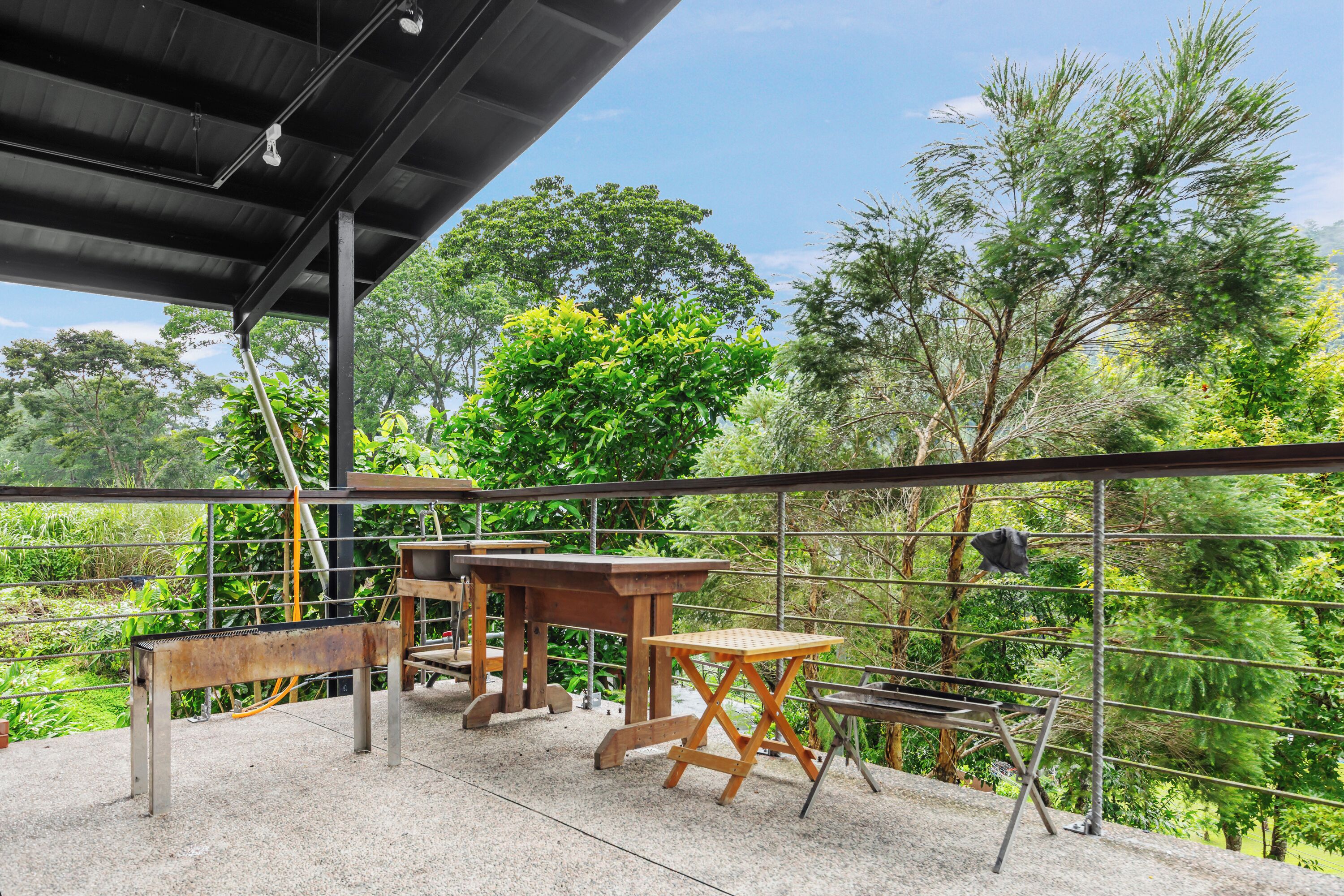 Un patio exterior con una mesa de madera y bancos, rodeado por una valla de metal y abundante vegetación.
