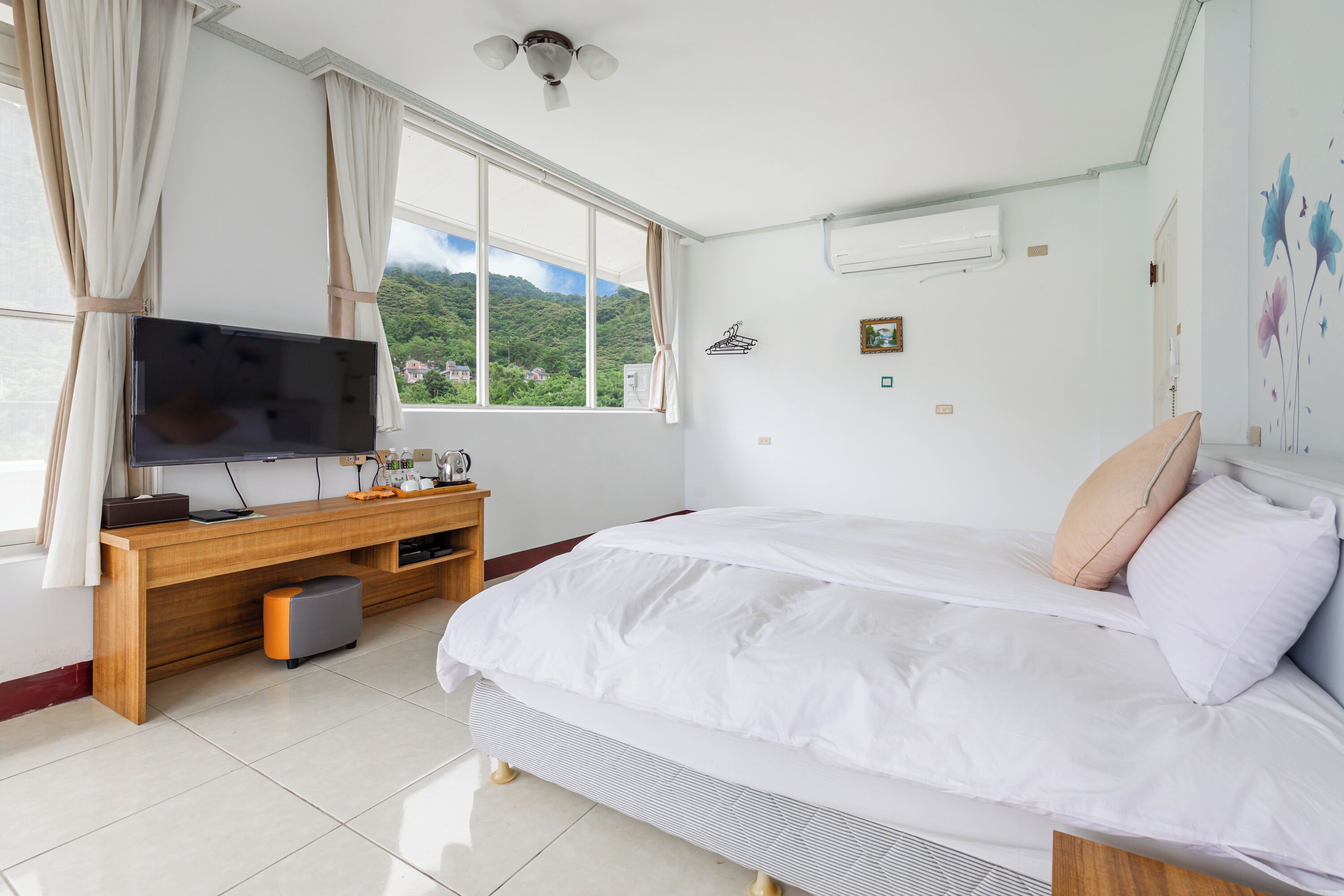 A bedroom with a bed, a TV on a wooden desk, a window with curtains, and a wall-mounted air conditioner.