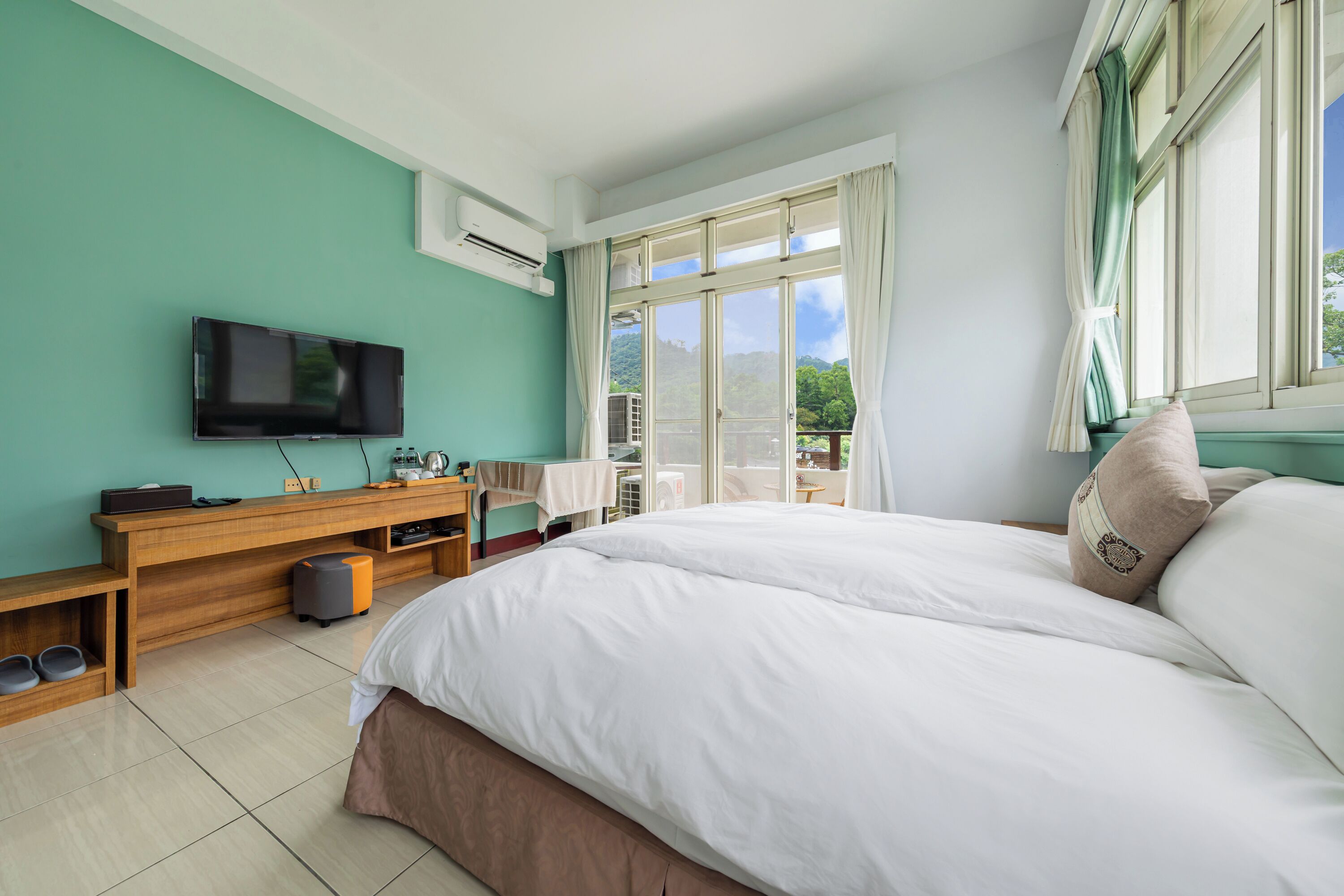 Un dormitorio con una cama grande, un televisor montado en la pared, un escritorio de madera y una ventana con cortinas.