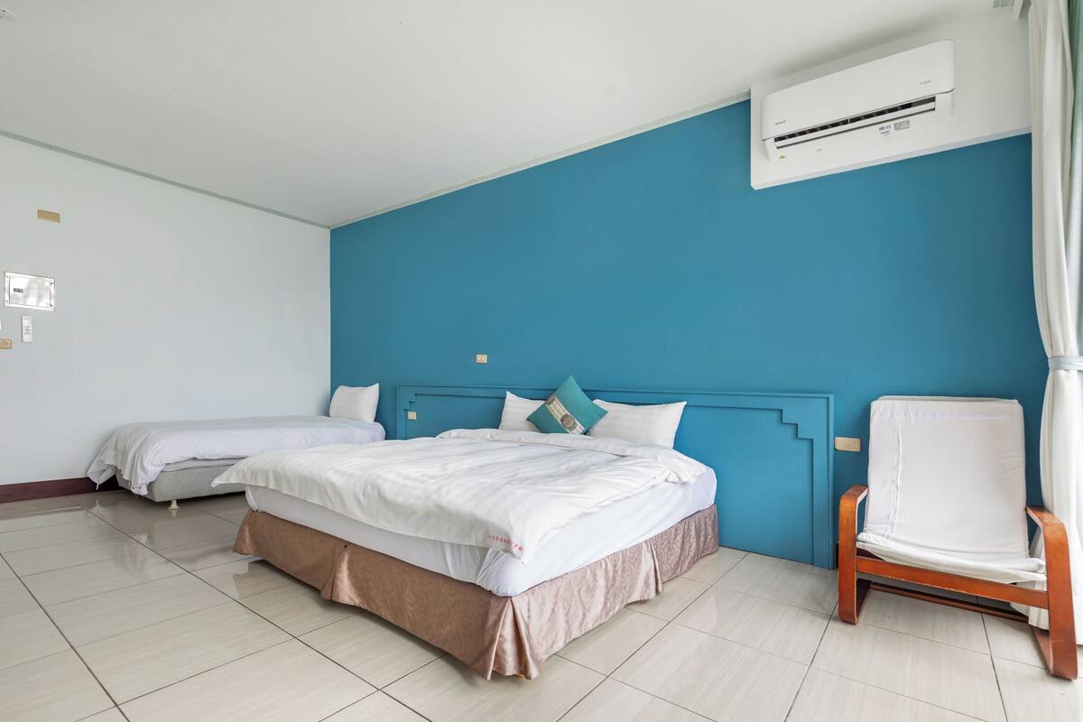 Une chambre d’hôtel avec deux lits, un mur d’accent bleu, une chaise en bois et un appareil de climatisation.
