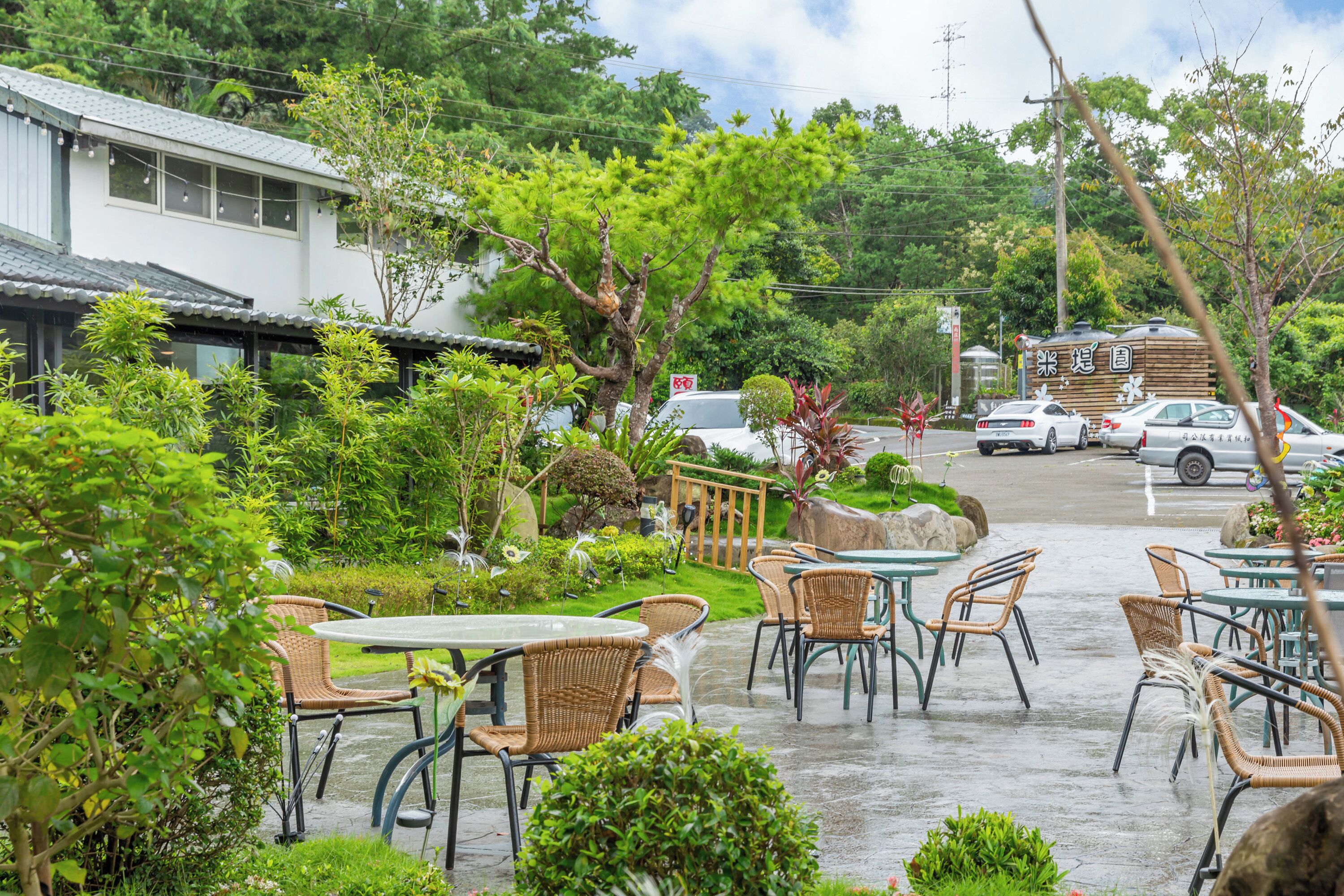 An outdoor cafe with wicker chairs and tables, surrounded by greenery and a building in the background.