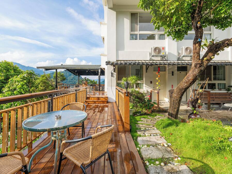 A wooden deck with a table and chairs, a stone pathway, and a building with a balcony.