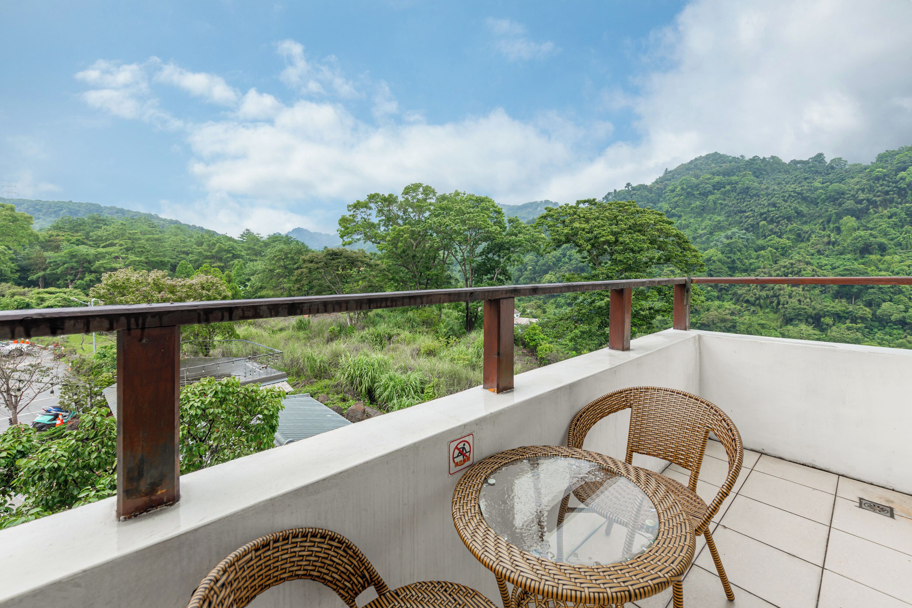 Ein Balkon mit Korbstühlen und einem Glastisch mit Blick auf eine üppige, grüne Landschaft und Berge in der Ferne.