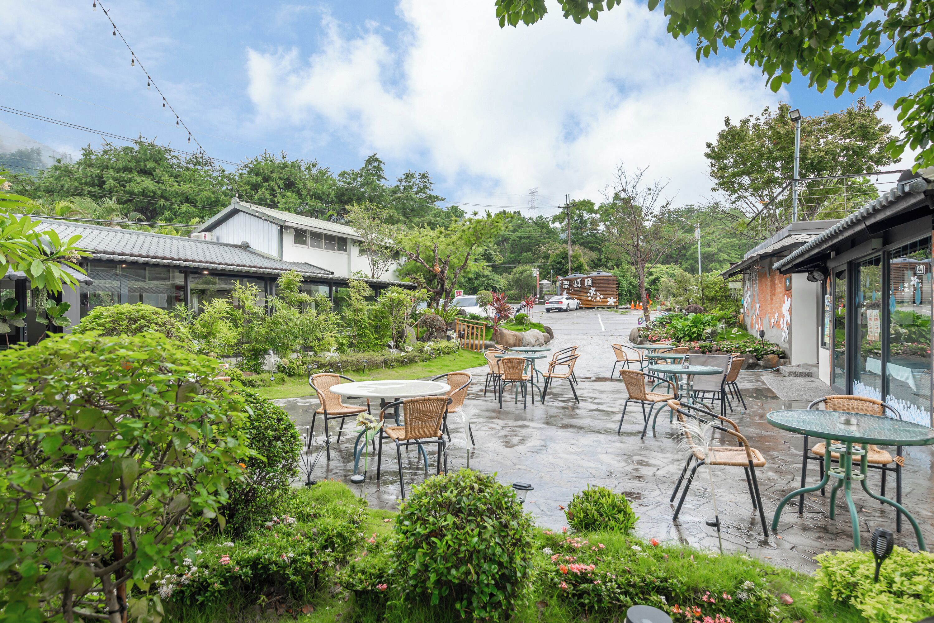 An outdoor cafe with tables and chairs, surrounded by lush greenery and traditional buildings.