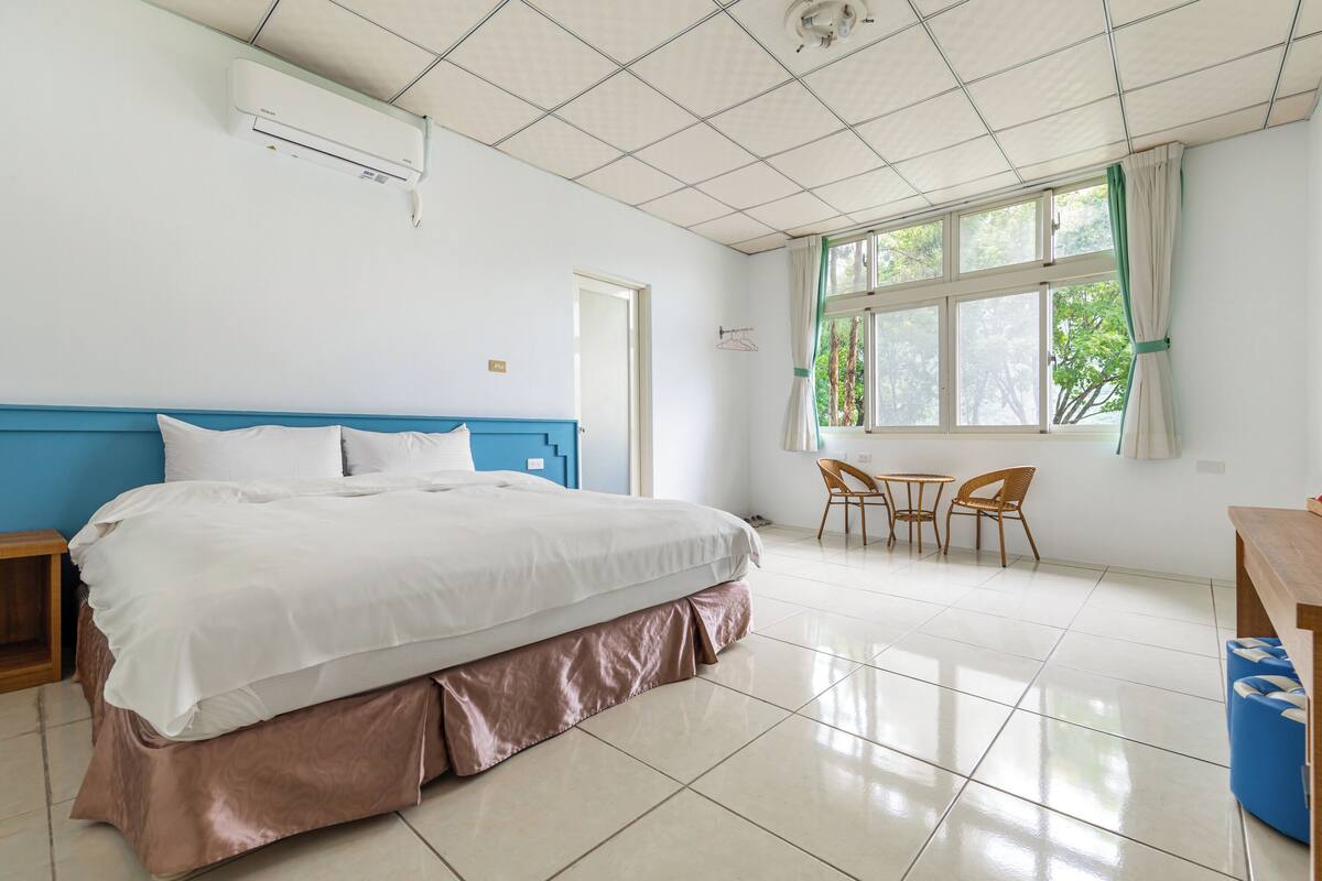 Une chambre d’hôtel avec un grand lit, une fenêtre avec des rideaux, une petite table et une chaise.