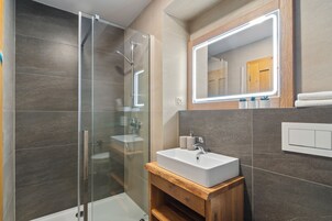 Un baño moderno con cabina de ducha de vidrio, tocador de madera con lavamanos y un espejo grande con iluminación integrada.
