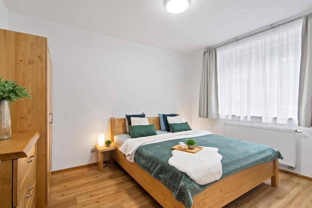 A bedroom with a wooden bed, a bedside table with a lamp, a window with curtains, and a wooden wardrobe.