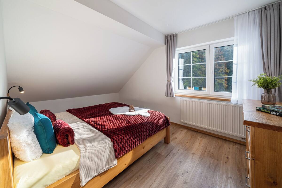 Un dormitorio con techo inclinado, una cama con manta roja, una lámpara de noche, un radiador, una ventana con cortinas y una planta pequeña en una mesita de noche.