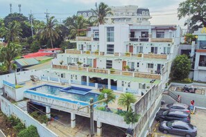 Un hotel de varios pisos con piscina en la azotea, balcones y un estacionamiento.
