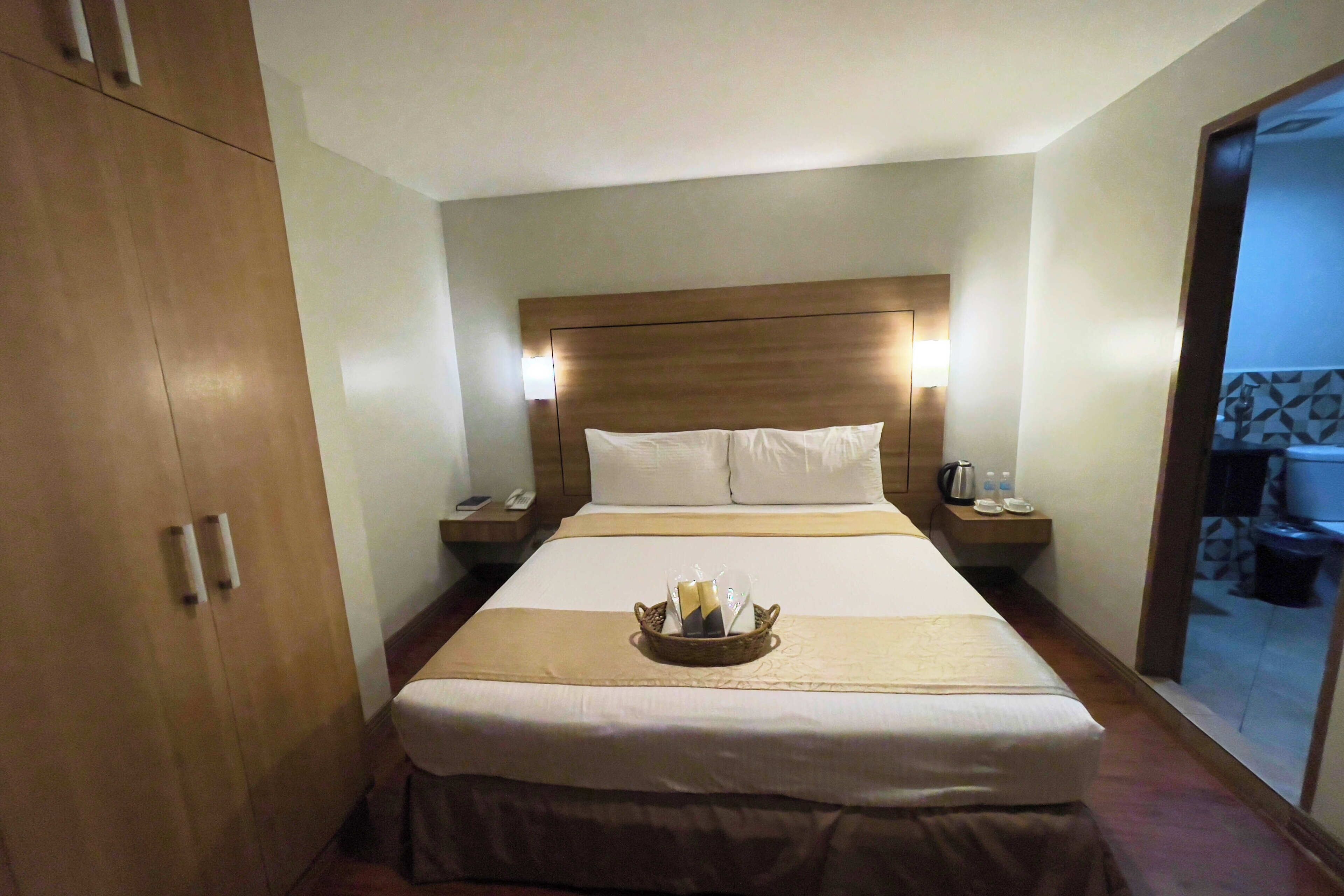 Una habitación de hotel con una cama grande, cabecera de madera, mesitas de noche con lámparas y una pequeña bandeja con artículos de aseo.