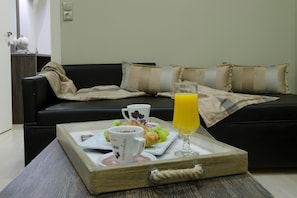 A breakfast tray with croissants, grapes, a glass of orange juice, and a coffee cup on a wooden tray in a living room with a black sofa and beige cushions.