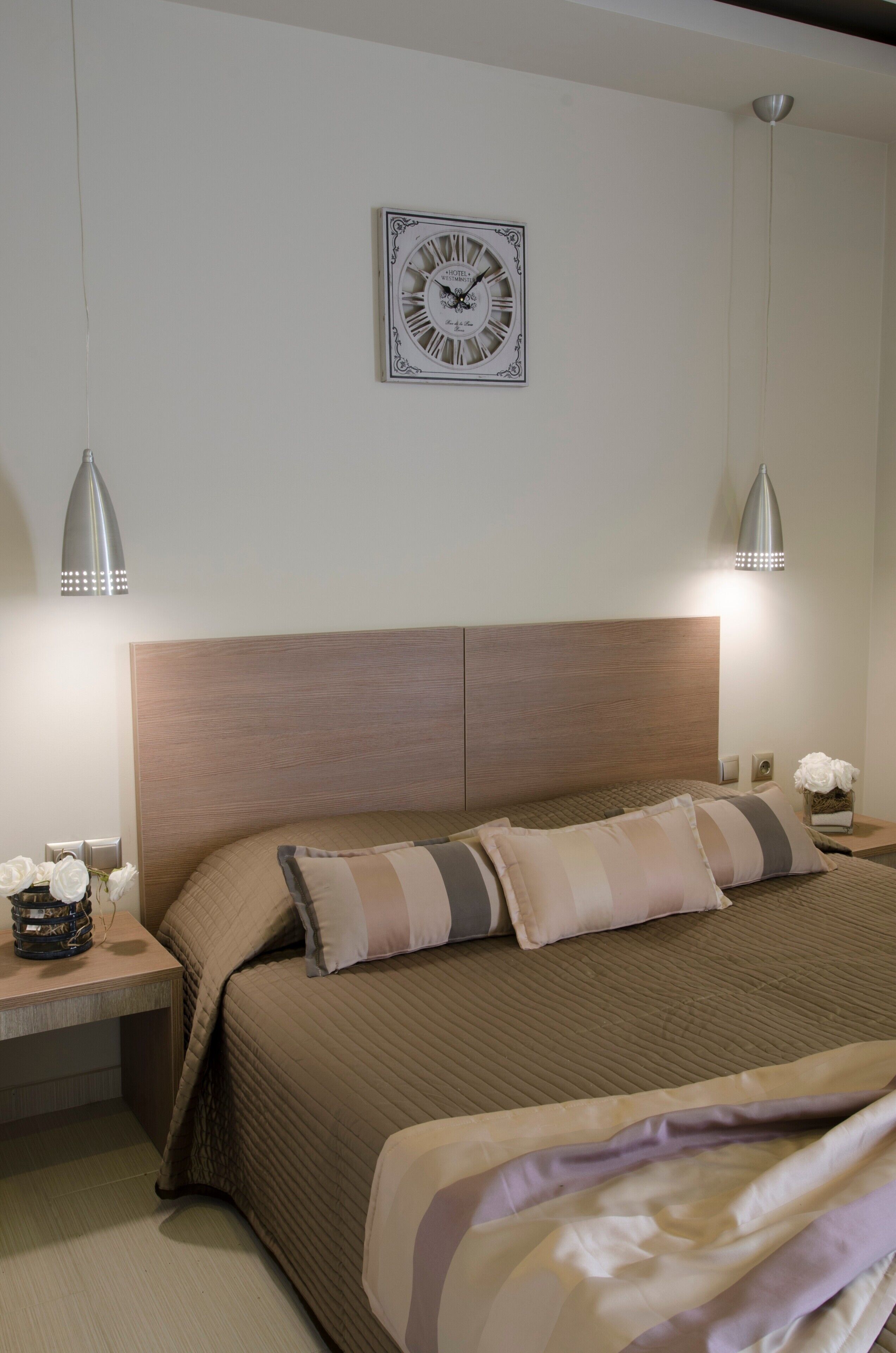 Une chambre à coucher avec une tête de lit en bois, un lit avec une literie à rayures, deux lampes de chevet, une horloge murale et une table de chevet ornée d’un vase de fleurs.