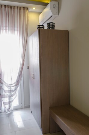 A room with a wardrobe, a bench, a window with curtains, and an air conditioning unit.