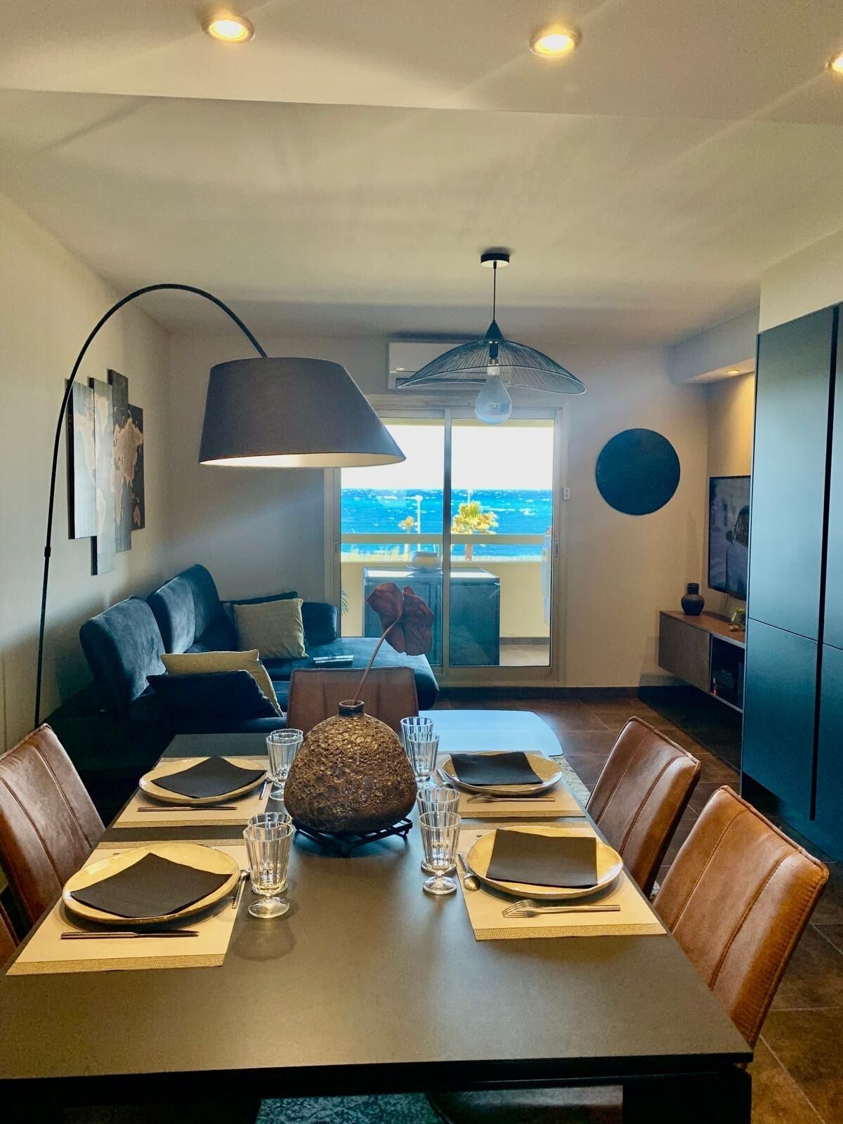 Un comedor moderno con una mesa dispuesta para cuatro, una lámpara colgante grande y vistas al mar a través de un ventanal.