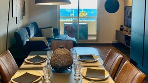 A modern dining area with a table set for four, a large pendant light, and a view of the sea through a large window.