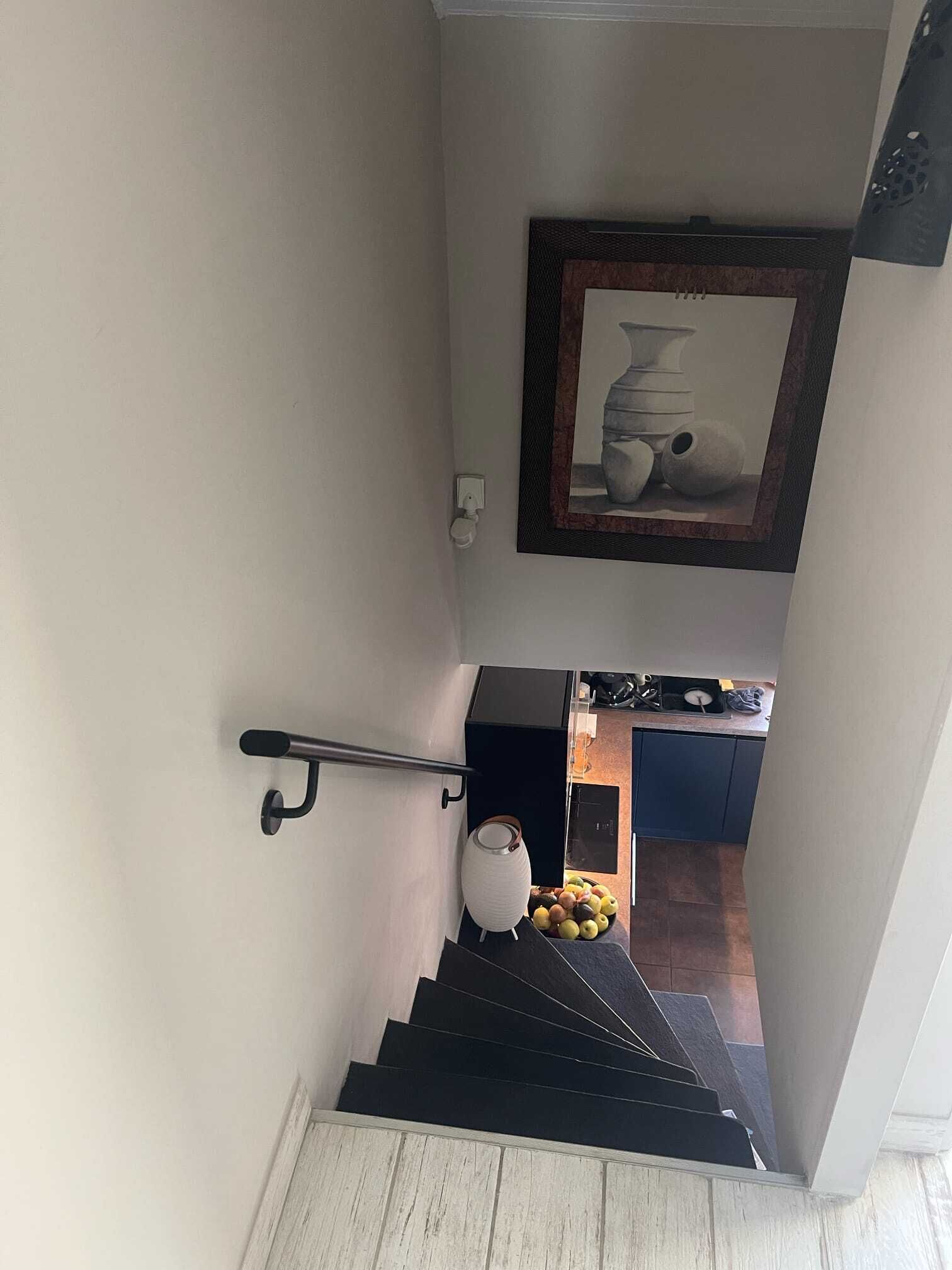 Una escalera con barandilla, una obra de arte enmarcada en la parte superior y una zona de cocina con un frutero a la vista.