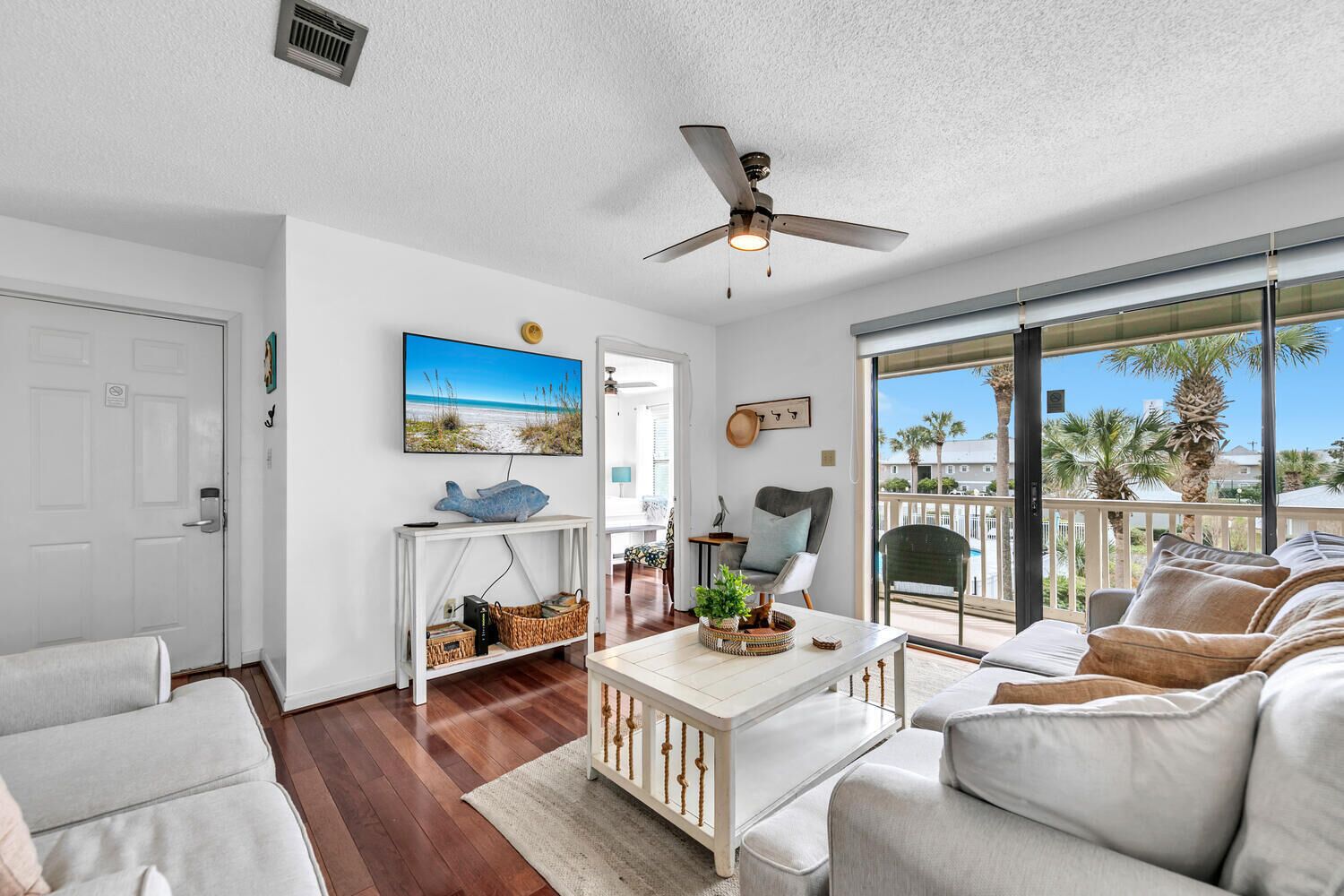 A spacious living room with a white ceiling fan, a large window with a view of palm trees, a white sofa, a coffee table, and a TV mounted on the wall.