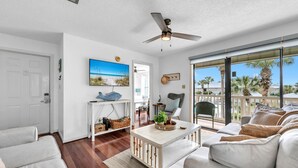 Ein geräumiges Wohnzimmer mit Deckenventilator, großem Fenster mit Blick auf Palmen, weißem Sofa, Couchtisch und Wand-Fernseher.