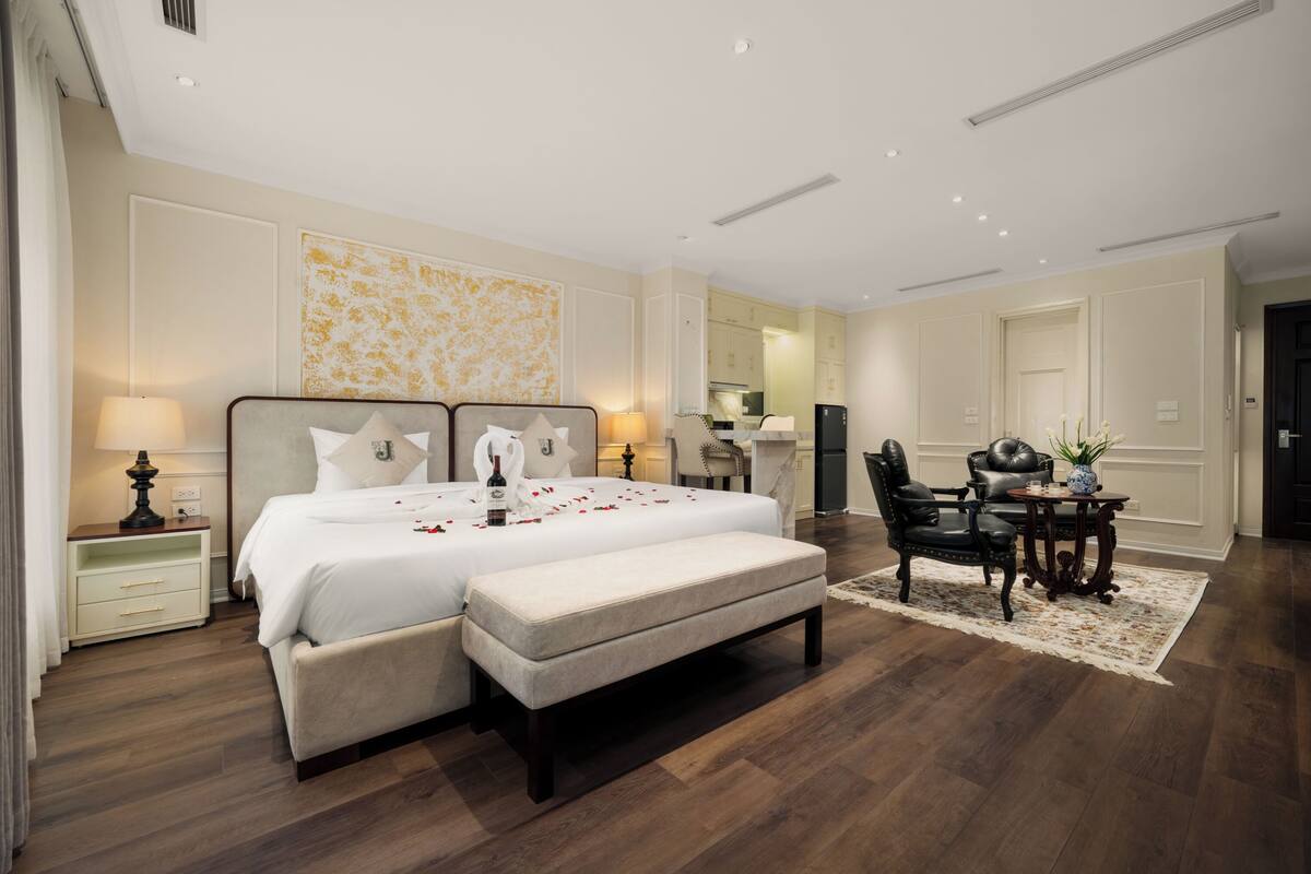 Un dormitorio amplio con una cama grande, mesitas de noche, un banco, una zona de comedor con mesa y sillas, y una cocina al fondo.