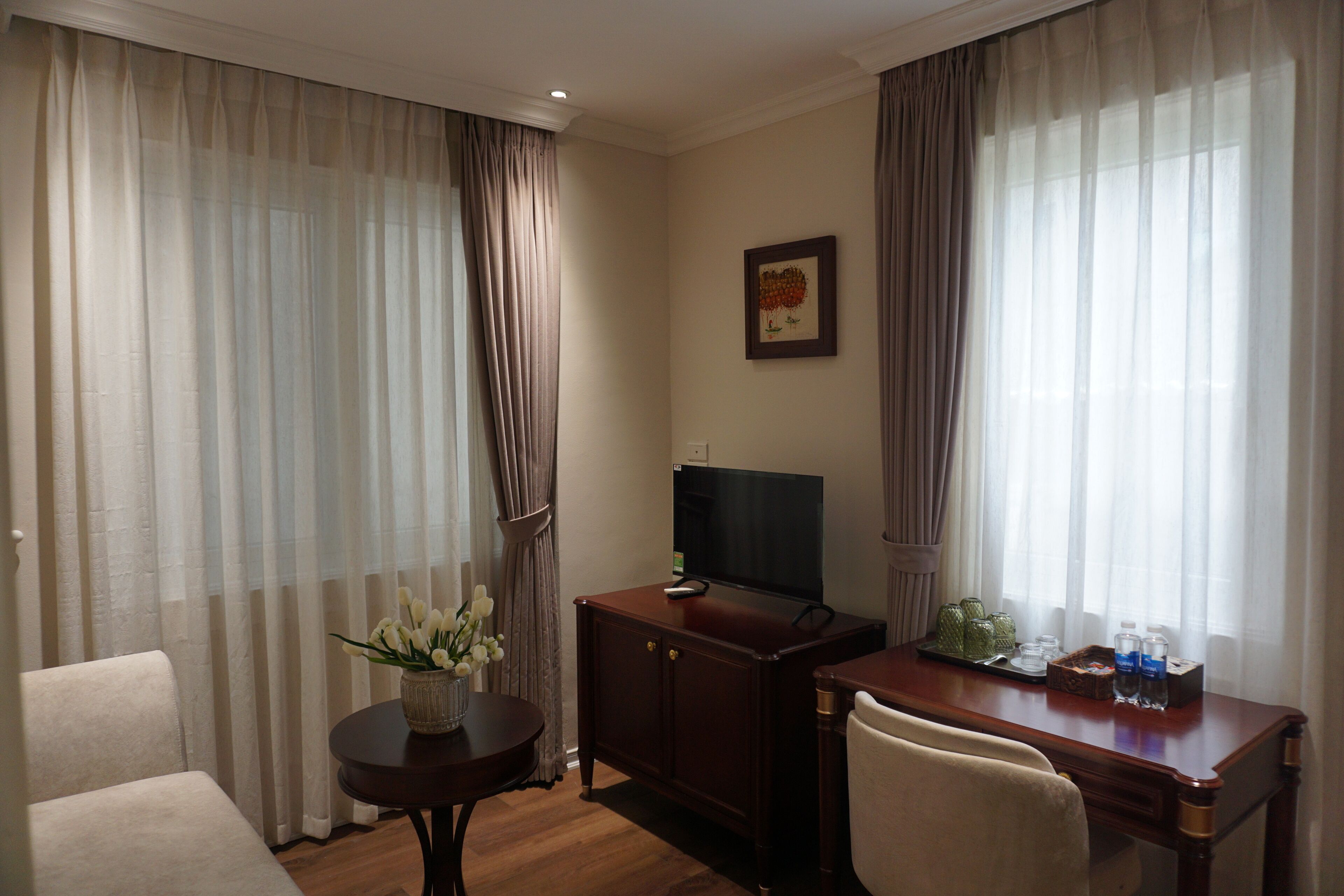A hotel room with a wooden desk, a television, a vase with flowers, and a framed picture on the wall.