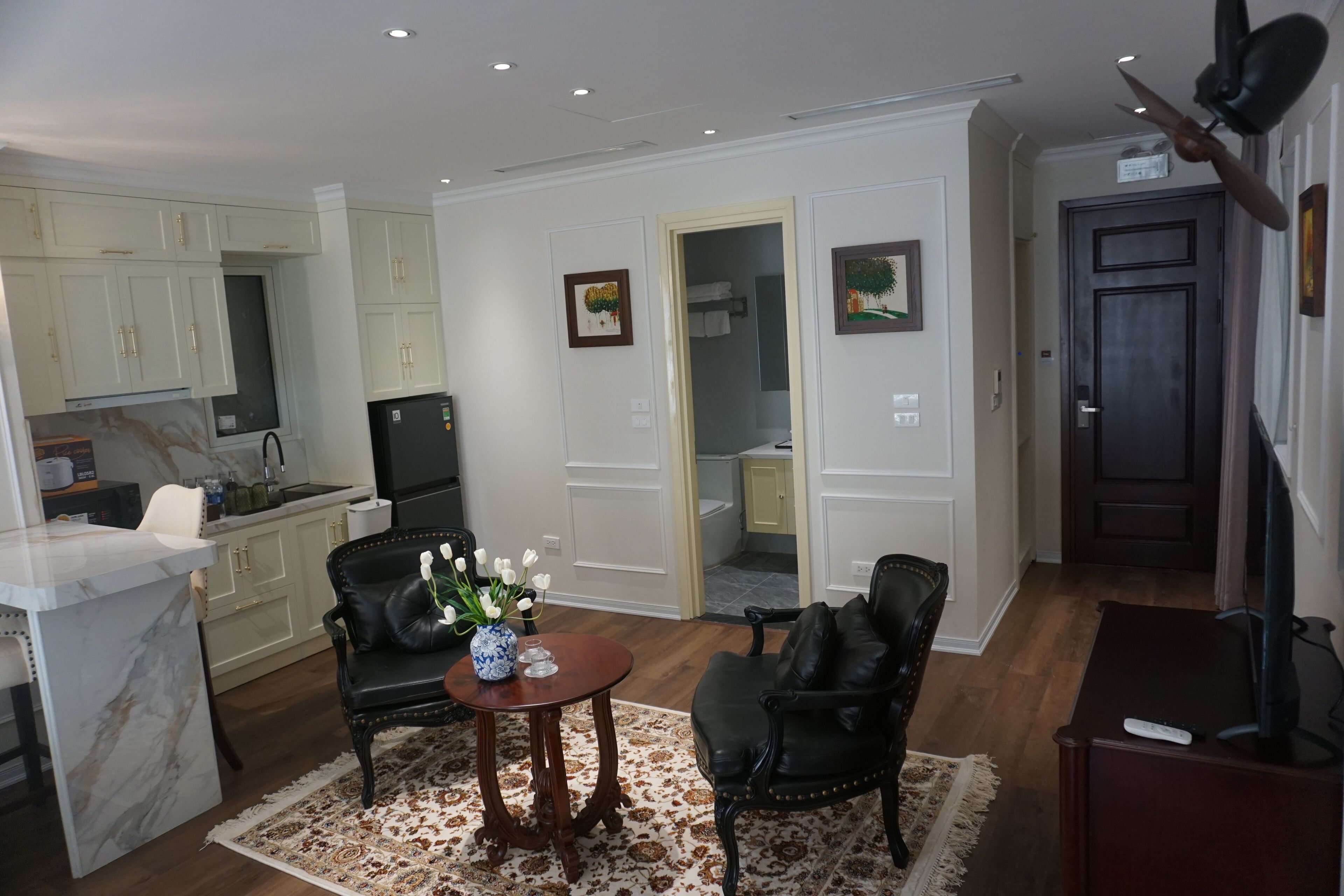 A modern living room with a marble kitchen island, black leather chairs, a small round table with a vase of flowers, and a television on a wooden stand.