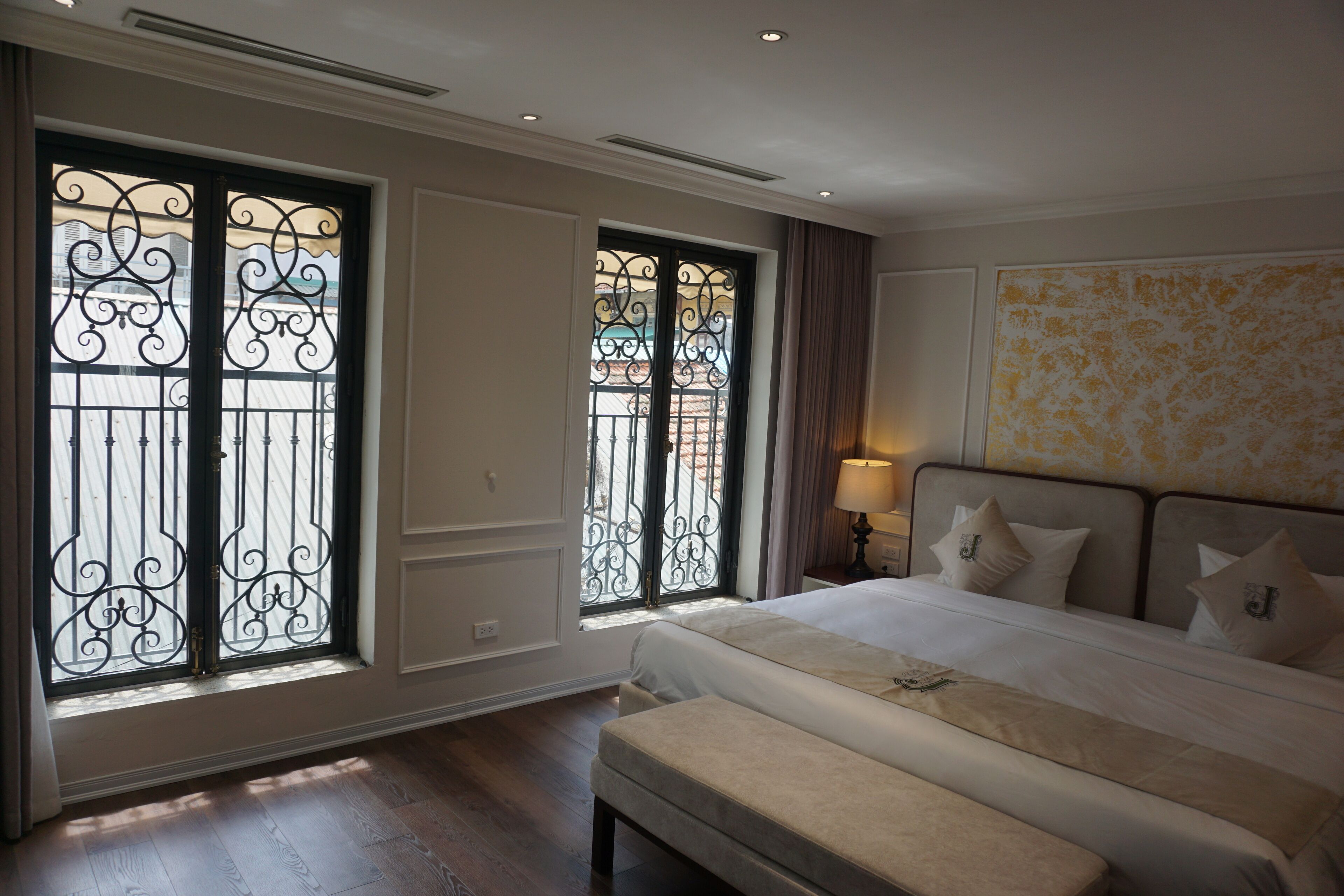 A hotel room with a large bed, a bench, and two windows with decorative iron grills.