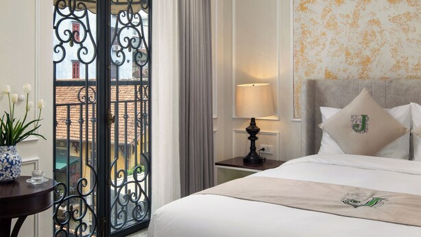 Quarto de hotel com uma cama grande, uma mesa de cabeceira com abajur, uma planta em vaso e vista para a varanda.