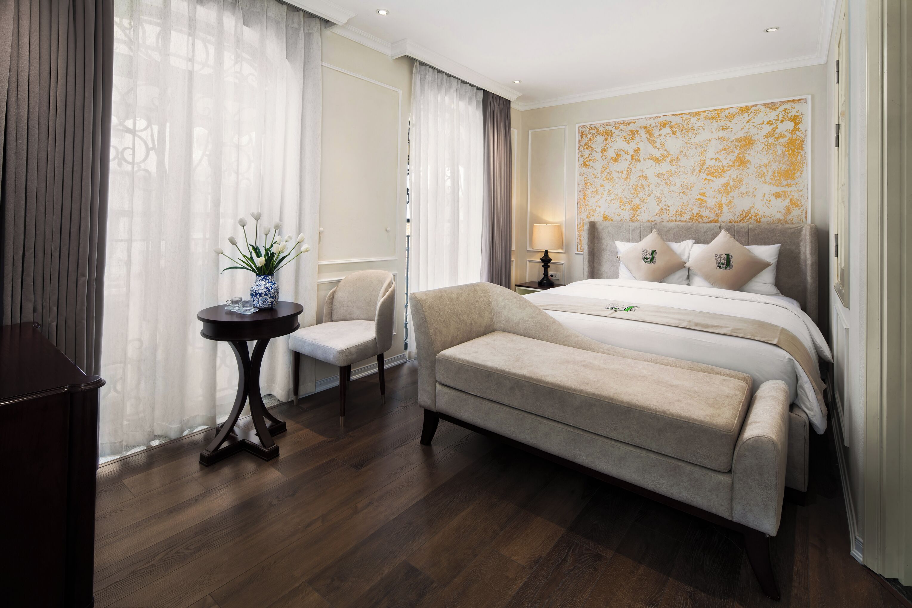 Una habitación de hotel con cama, mesita de noche, una silla, una pequeña mesa con un florero y un ventanal grande con cortinas.