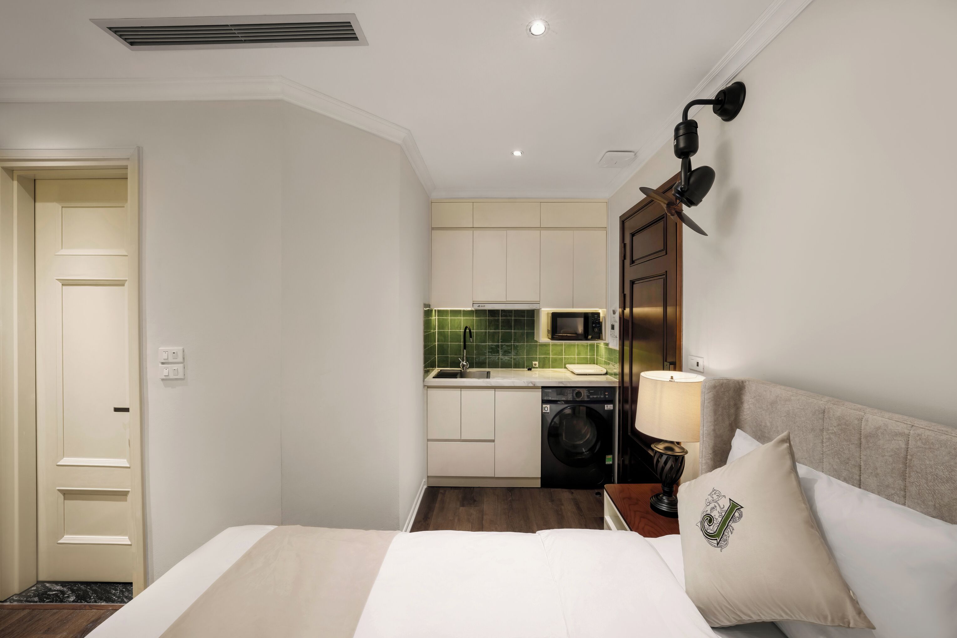 A compact hotel room with a bed, a bedside table with a lamp, a small kitchen area with a washing machine, and a door leading to another room.