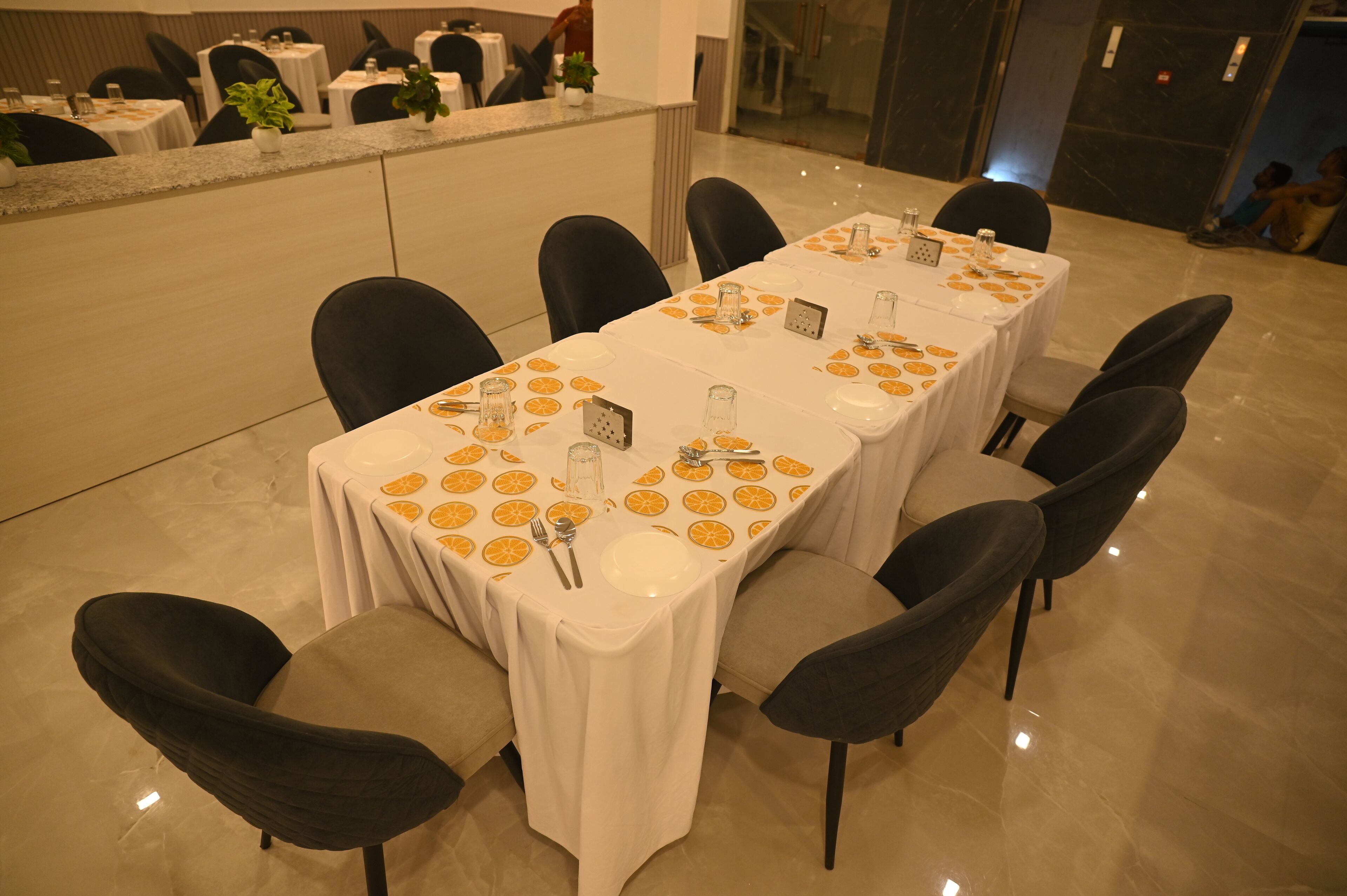 A long dining table with a white tablecloth and orange fruit pattern, surrounded by black chairs in a modern hotel setting.