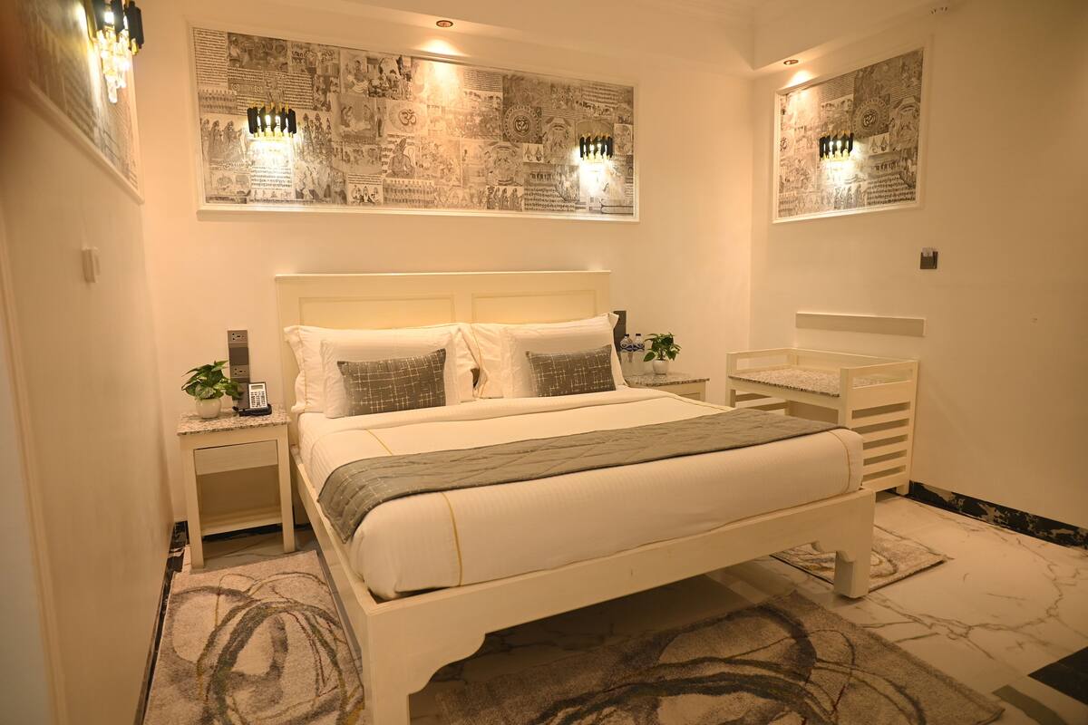 A bedroom with a large bed, a bedside table with a lamp, a nightstand, a chair, and a wall-mounted artwork.