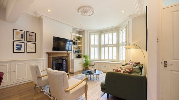 A living room with a fireplace, a television, a bookshelf, and a window with white shutters. - The Battersea Park Townhouse (London)