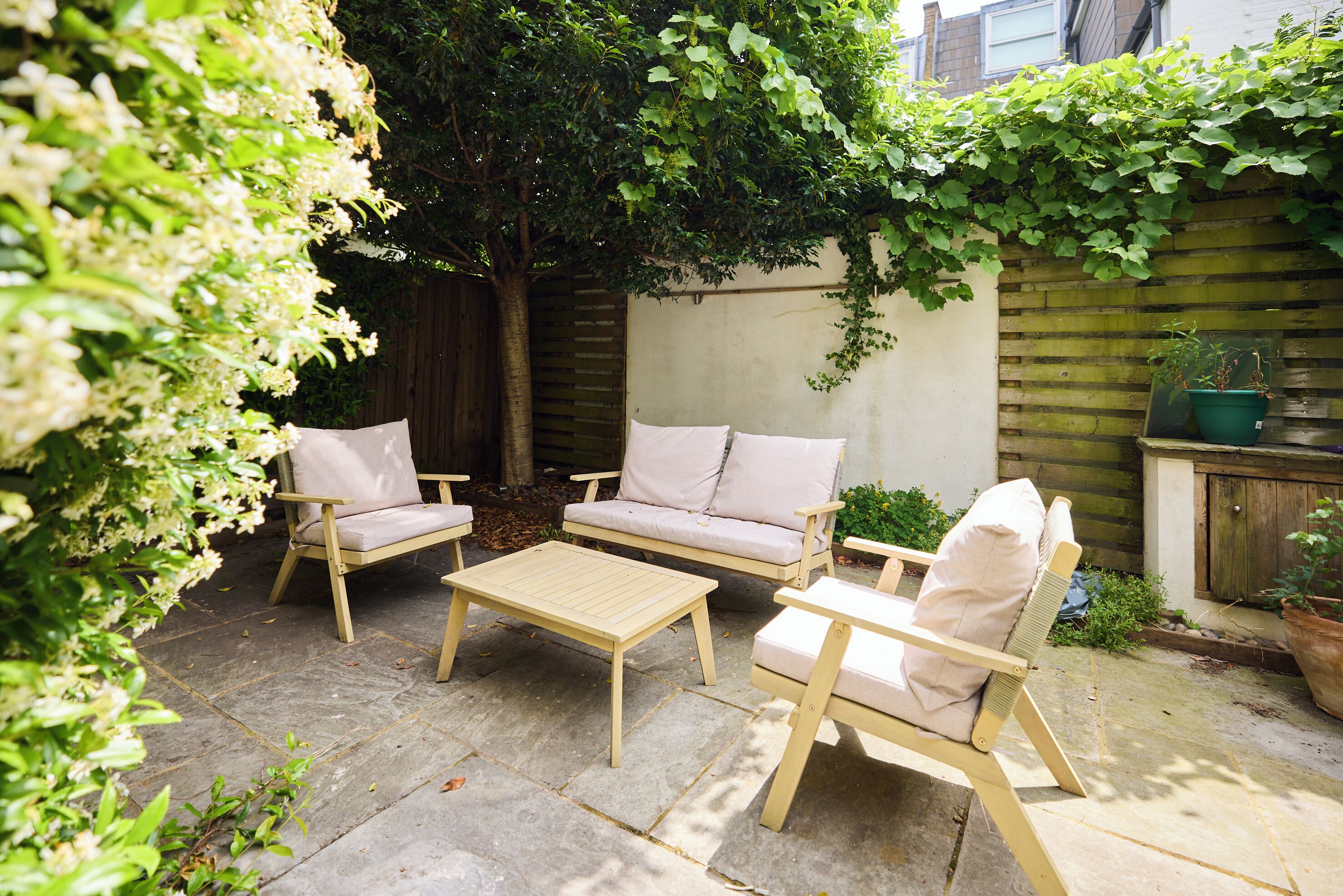 A patio with a wooden table and two cushioned chairs, surrounded by greenery and a wooden fence.