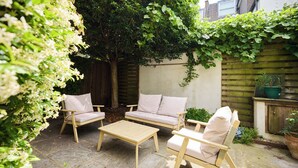 A patio with a wooden table and two cushioned chairs, surrounded by greenery and a wooden fence. - The Battersea Park Townhouse (London)