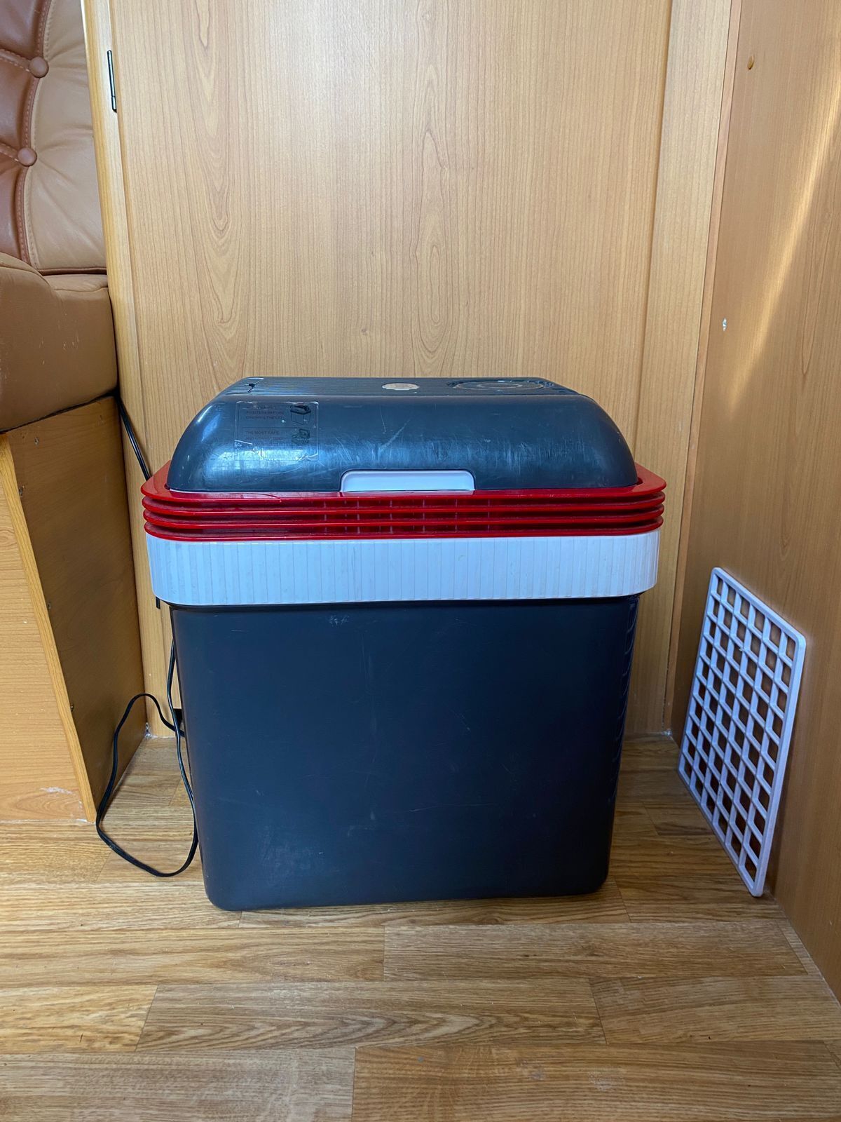 A portable cooler with a red lid and a white base, placed on a wooden floor next to a white vent.