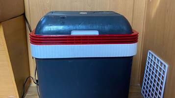 A portable cooler with a red lid and a white base, placed on a wooden floor next to a white vent.