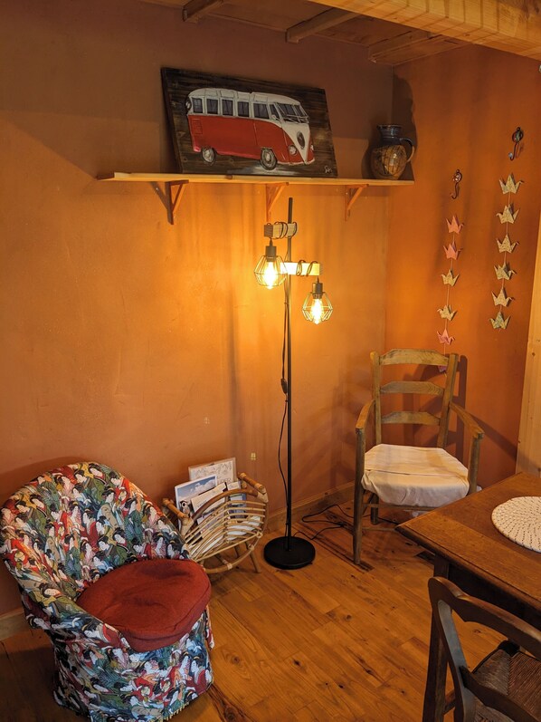 Ein gemütliches Zimmer mit Holzboden, einem roten und weißen Vintage-Autobild an der Wand, einer Stehlampe mit drei Glühbirnen, einem Holzstuhl, einem kleinen Tisch und einem farbenfrohen Kissen auf einem Stuhl.
