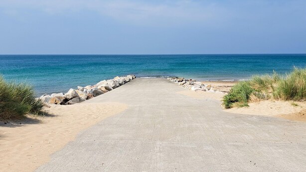 Een geplaveid pad dat naar een strand leidt, met een rotsachtige zeedijk en helderblauw water.