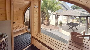 A wooden sauna with a bench, a wooden door, and a view of an outdoor patio with a pergola, plants, and a wheelbarrow.