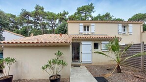 Una casa de dos pisos con techo de tejas, puerta principal y un pequeño jardín con plantas en macetas.